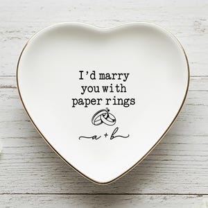 May include: A heart-shaped white ceramic trinket dish with a gold rim. The dish features the text "I'd marry you with paper rings" and a graphic of two rings. The bottom of the dish has the initials "a + b".