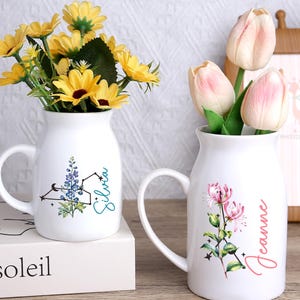 Peut inclure: Deux vases en céramique blancs avec des poignées, chacun contenant des fleurs. L'un a des fleurs jaunes et un motif floral bleu avec le nom "Silvia". L'autre a des fleurs roses et le nom "Jeanne". Un cadre photo en bois est en arrière-plan.