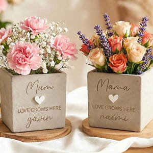 Op de afbeelding: Twee vierkante, grijze betonnen plantenbakken met bloemen. Eén heeft roze anjers en gipskruid, de andere rozen en lavendel. Elke plantenbak heeft een hart en de woorden "Mum" en "Mama" met namen eronder. De plantenbakken staan op houten onderzetters.