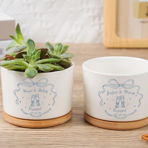 Puede incluir: Dos macetas de cerámica blanca con un diseño azul claro y bases de bambú. Una maceta contiene una suculenta. El diseño incluye texto y copas de champán, con los nombres "Hugo & Betsy" y "Jafar & Merry".