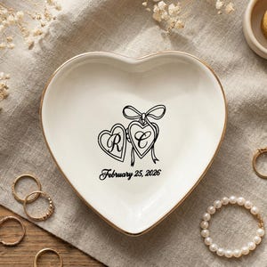 Puede incluir: Un plato de cerámica blanco en forma de corazón con borde dorado, con dos diseños de corazón conectados con iniciales y la fecha 25 de febrero de 2026. Rodeado de anillos, una pulsera de perlas y una vela encendida.