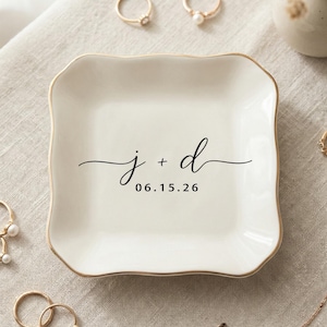 May include: A square, off-white ceramic ring dish with a gold rim and the inscription "j + d" and the date "06.15.26" in black script. Several gold rings and a necklace are arranged around the dish.