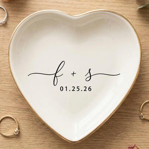 May include: A heart-shaped, white ceramic ring dish with a gold rim. The dish is inscribed with the letters "f + s" and the date "01.25.26" in black script. Several rings are scattered around the dish on a wooden surface.