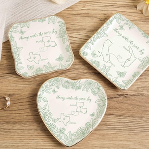 May include: Three decorative trinket dishes in square, diamond, and heart shapes. Each dish features a floral border, state outlines, names, and the phrase "Always under the same sky" in green. Gold trim edges the dishes.