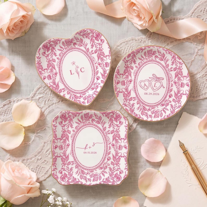 May include: Three ceramic trinket dishes with pink floral patterns and gold rims. One heart-shaped dish features the letters L C. A round dish displays two hearts with A L and 08.29.2026. A square dish shows f + s and 09.15.2026 within oval frames.