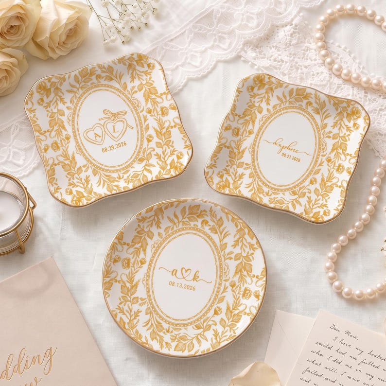 May include: Three white ceramic dishes with scalloped edges and gold floral borders. Two square dishes feature oval centers with heart motifs and dates 08.29.2026 and 08.13.2026. One square dish features an oval center with the name Daphne and 08.21.2026.