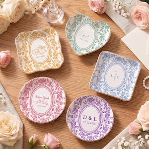May include: Five ceramic trinket dishes in square, heart, and round shapes, featuring floral borders and personalized text including initials and dates. The dishes are colored in gold, green, blue, pink, and purple, each with a central oval design.