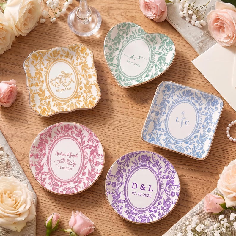 May include: Five ceramic trinket dishes in square, heart, and round shapes, featuring floral borders and personalized text including initials and dates. The dishes are colored in gold, green, blue, pink, and purple, each with a central oval design.