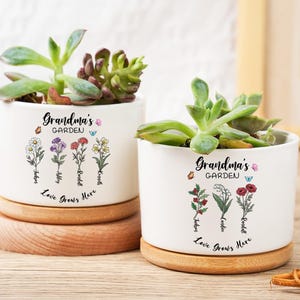 Könnte beinhalten: Zwei weiße Keramik-Pflanzgefäße mit Sukkulenten, jeweils auf einem Holzsockel. Die Pflanzgefäße sind mit floralen Illustrationen und dem Text "Grandma's Garden" und "Love Grows Here" verziert. Eine charmante Ergänzung für jedes Zuhause.