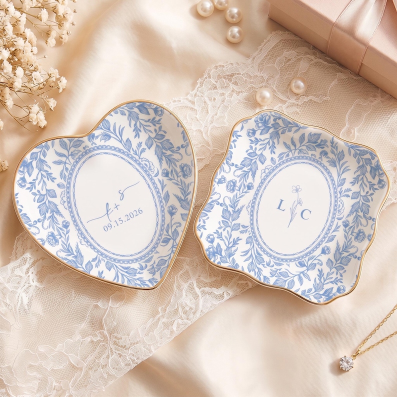 May include: Two white ceramic trinket dishes with gold-toned rims and blue floral patterns. One dish is heart-shaped with the text 'f + s' and '09.15.2026'. The other is square with scalloped edges and the letters 'L C' featuring a small flower icon.