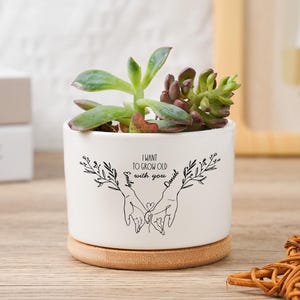 Op de afbeelding: Een witte keramische plantenpot met een vetplant. De pot heeft de tekst "I want to grow old with you" en de namen "Emily" en "Daniel". Hij staat op een ronde houten basis.