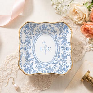 May include: A square ceramic trinket dish with scalloped edges and a gold-toned rim. The dish features a white center with a blue floral oval border, the letters L and C, and a small flower illustration. The exterior border has a blue floral pattern.