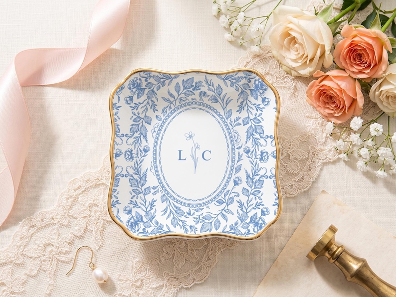 May include: A square ceramic trinket dish with scalloped edges and a gold-toned rim. The dish features a white center with a blue floral oval border, the letters L and C, and a small flower illustration. The exterior border has a blue floral pattern.