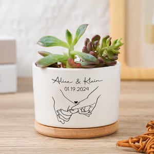 May include: A white ceramic plant pot with a succulent, inscribed with "Alice & Klein" and the date "01.19.2024", and a line drawing of two hands holding pinky fingers. The pot rests on a round wooden base.