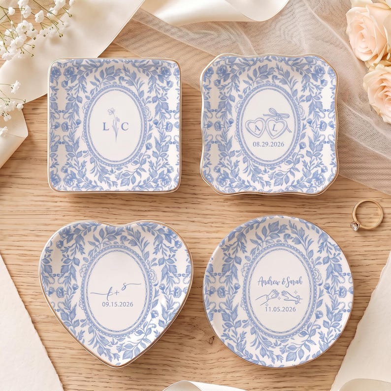 May include: Four ceramic trinket dishes with blue and white floral patterns and gold-toned rims. The dishes feature personalized text including initials L C, A L with 08.29.2026, t + s with 09.15.2026, and Andrew & Sarah with 11.05.2026, plus heart and hand icons.