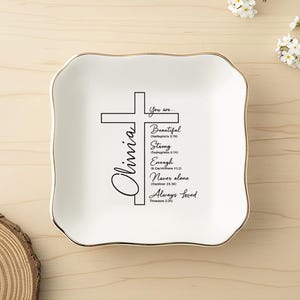 May include: A square white ceramic trinket dish with a gold-trimmed scalloped edge. The center features a black cross graphic with the name Olivia and the text: You are Beautiful, Strong, Enough, Never alone, Always Loved, with Bible verses.