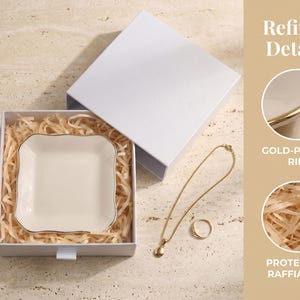 May include: A square white ceramic trinket dish with a gold-plated rim, nestled in a white gift box with protective raffia fill. The set includes a gold-tone chain necklace with a small pendant and a simple gold-tone ring, presented for display.