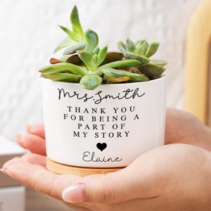 May include: A white ceramic planter with a succulent plant, held in hands. The planter has the text "Mrs. Smith, Thank you for being a part of my story" with a heart and the name "Elaine". The planter sits on a wooden base.