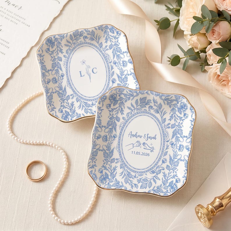 May include: Two square ceramic trinket dishes with scalloped edges and gold trim, featuring a blue and white floral pattern. One dish displays the letters L C, while the other shows the text Andrew & Sarah with a hand illustration and 11.05.2026.