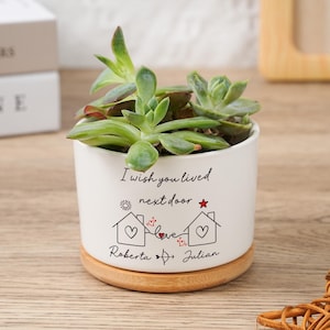 Può includere: Un vaso di ceramica bianco con una pianta succulenta. Il vaso ha una base in legno e presenta il testo "I wish you lived next door" con illustrazioni di case e i nomi Roberta e Julian. La succulenta ha foglie verdi e rosse.