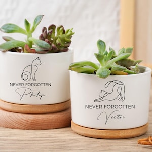 May include: Two white ceramic plant pots with succulents, each featuring a black line drawing of a cat and the words "NEVER FORGOTTEN" with a name below. The pots sit on wooden saucers.