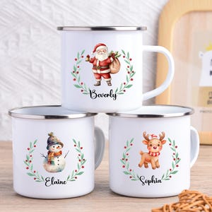 Könnte beinhalten: Drei weiße Emaille-Tassen mit silbernen Rändern, jede mit einem anderen Weihnachtscharakter: Weihnachtsmann, Schneemann und Rentier. Jede Tasse hat einen Namen unter dem Charakter aufgedruckt und ist von einem grünen Kranz mit roten Beeren umgeben.