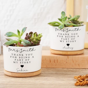May include: Two white ceramic planters with succulents, each on a wooden base. The planters have the text "Mrs. Smith, Thank you for being a part of my story" with a name below. One has "Sabina" and the other "Audrey".