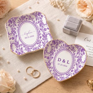 May include: Two ceramic trinket dishes with purple floral patterns and gold-trimmed edges. One square dish features the name Daphne and the date 08.21.2026. The heart-shaped dish displays the initials D & L and the date 07.23.2026 in the center.