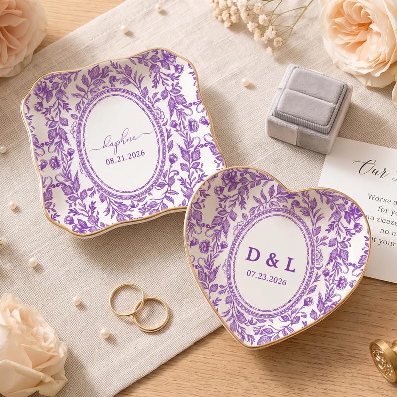 May include: Two ceramic trinket dishes with purple floral patterns and gold-trimmed edges. One square dish features the name Daphne and the date 08.21.2026. The heart-shaped dish displays the initials D & L and the date 07.23.2026 in the center.