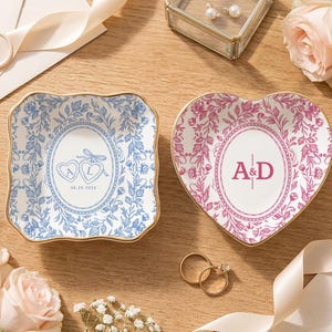 May include: Two ceramic trinket dishes with gold-toned rims. One square dish features blue floral patterns and two hearts with the letters A and L and the date 08.29.2026. The other heart-shaped dish features pink floral patterns and the letters A&D.
