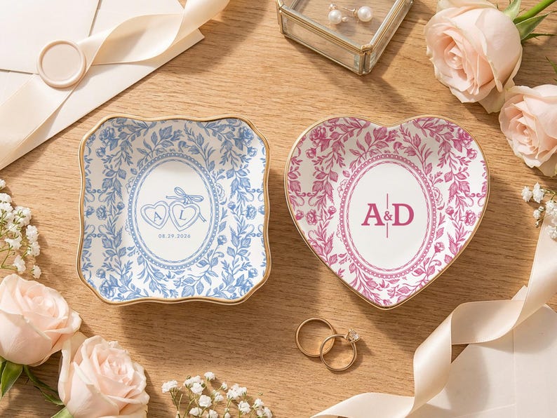 May include: Two ceramic trinket dishes with gold-toned rims. One square dish features blue floral patterns and two hearts with the letters A and L and the date 08.29.2026. The other heart-shaped dish features pink floral patterns and the letters A&D.