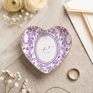 May include: A heart-shaped ceramic trinket dish featuring a purple floral pattern and a central oval frame. The center contains the script letters f + s and the date 09.15.2026. The dish has a thin gold-colored rim along the outer edge.