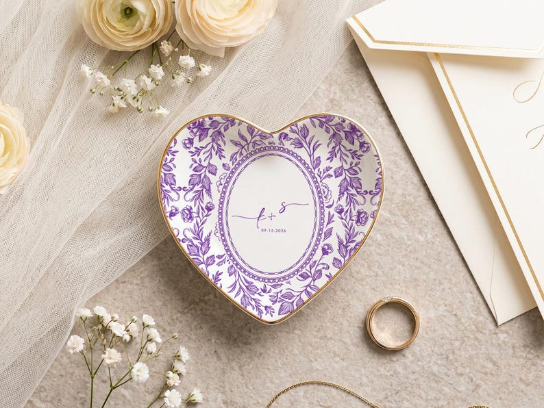 May include: A heart-shaped ceramic trinket dish featuring a purple floral pattern and a central oval frame. The center contains the script letters f + s and the date 09.15.2026. The dish has a thin gold-colored rim along the outer edge.