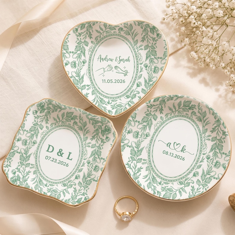 May include: Three ceramic trinket dishes with gold-toned rims and green botanical patterns. One heart-shaped dish reads Andrew & Sarah 11.05.2026, one square dish reads D & L 07.23.2026, and one round dish reads a heart k 08.13.2026. A gold ring sits nearby.