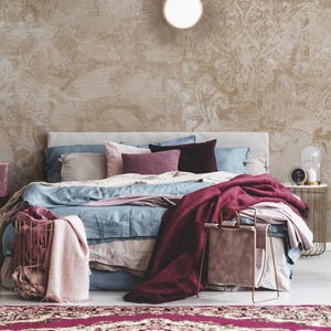 May include: A bedroom scene featuring a bed with blue and pink bedding, a burgundy throw, and decorative pillows. A gold wire basket holds a pink blanket. The room has a patterned wallpaper and a gold side table.