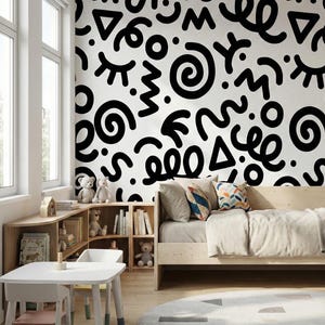 May include: A child's bedroom with a white wall featuring a black abstract pattern. The room includes a wooden bed, a small white table with two chairs, a bookshelf, and a round rug with geometric shapes. The room is well-lit by natural light.