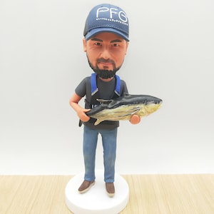 May include: A figurine of a man wearing a blue cap with "PFG" on it, a black shirt, blue jeans, and brown shoes, holding a fish. The figurine is on a white base. The fish is black and yellow.