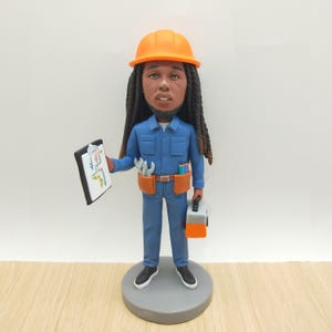 Op de afbeelding: Een beeldje van een persoon in blauwe werkkleding, een oranje helm en zwarte schoenen. De figuur houdt een klembord vast met een kleurrijk diagram en een grijze en oranje gereedschapskist. Een bruine gereedschapsriem met gereedschap zit om de taille.