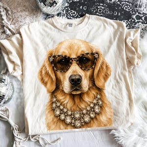 May include: A cream-colored t-shirt featuring a graphic print of a golden retriever wearing leopard print glasses and a large, multi-layered crystal statement necklace. The dog is centered on the front of the shirt, which has a crew neckline.
