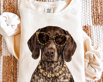 May include: A cream-coloured crewneck sweatshirt featuring a graphic print of a brown and white spotted dog wearing leopard print glasses and a chunky gold-toned beaded necklace. The interior neck label reads Comfort Colors in black text.