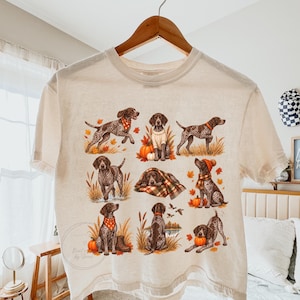 May include: A cream-colored short-sleeve t-shirt featuring a graphic print of nine German Shorthaired Pointer dogs in various autumn-themed poses, including wearing scarves and sweaters, surrounded by pumpkins, fall leaves, and tall grass.