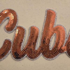 May include: A rose gold sequined iron-on patch with the word "Cubs" in a cursive font.
