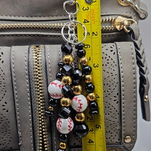 May include: A baseball-themed keychain charm with a silver clasp, featuring a beaded design of black, gold, and white baseballs with red stitching. The charm is approximately 3 inches long and is attached to a gray handbag.