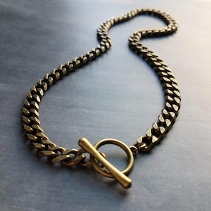 May include: A close-up of a gold-colored chain necklace with a toggle clasp. The chain is made of thick, interlocking links. The toggle clasp is a circular ring with a bar that fits through it. The necklace is on a dark gray surface.