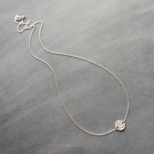 May include: A fine silver necklace featuring a hammered disc pendant. The pendant, with a textured surface, hangs from a delicate chain. The necklace is presented on a grey background, highlighting its minimalist style and understated gleam.