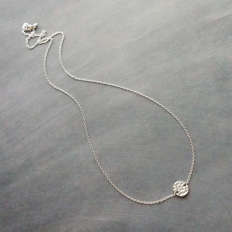 May include: A fine silver necklace featuring a hammered disc pendant. The pendant, with a textured surface, hangs from a delicate chain. The necklace is presented on a grey background, highlighting its minimalist style and understated gleam.