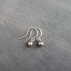 May include: A pair of small, silver-colored earrings. Each earring features a curved hook and a small, round bead. The earrings are displayed against a gray background, highlighting their simple design and metallic sheen.