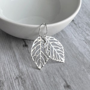 May include: Silver leaf earrings with intricate cut-out details. The earrings feature a delicate leaf design, with visible veins and a hook closure. The leaves are positioned in a layered arrangement, creating a three-dimensional effect. The earrings are displayed against a neutral background.