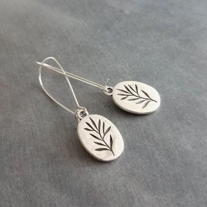 May include: Silver-toned dangle earrings. Each earring features an oval charm with a cut-out leaf design. The earrings have a simple, elegant design, perfect for everyday wear. The background is a solid gray.