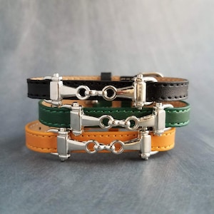 Op de afbeelding: Drie leren armbanden in zwart, groen en oranje, op elkaar gestapeld. Elke armband heeft een zilverkleurig metalen paardengebitontwerp en een gespsluiting. De armbanden zijn op elkaar gestapeld.